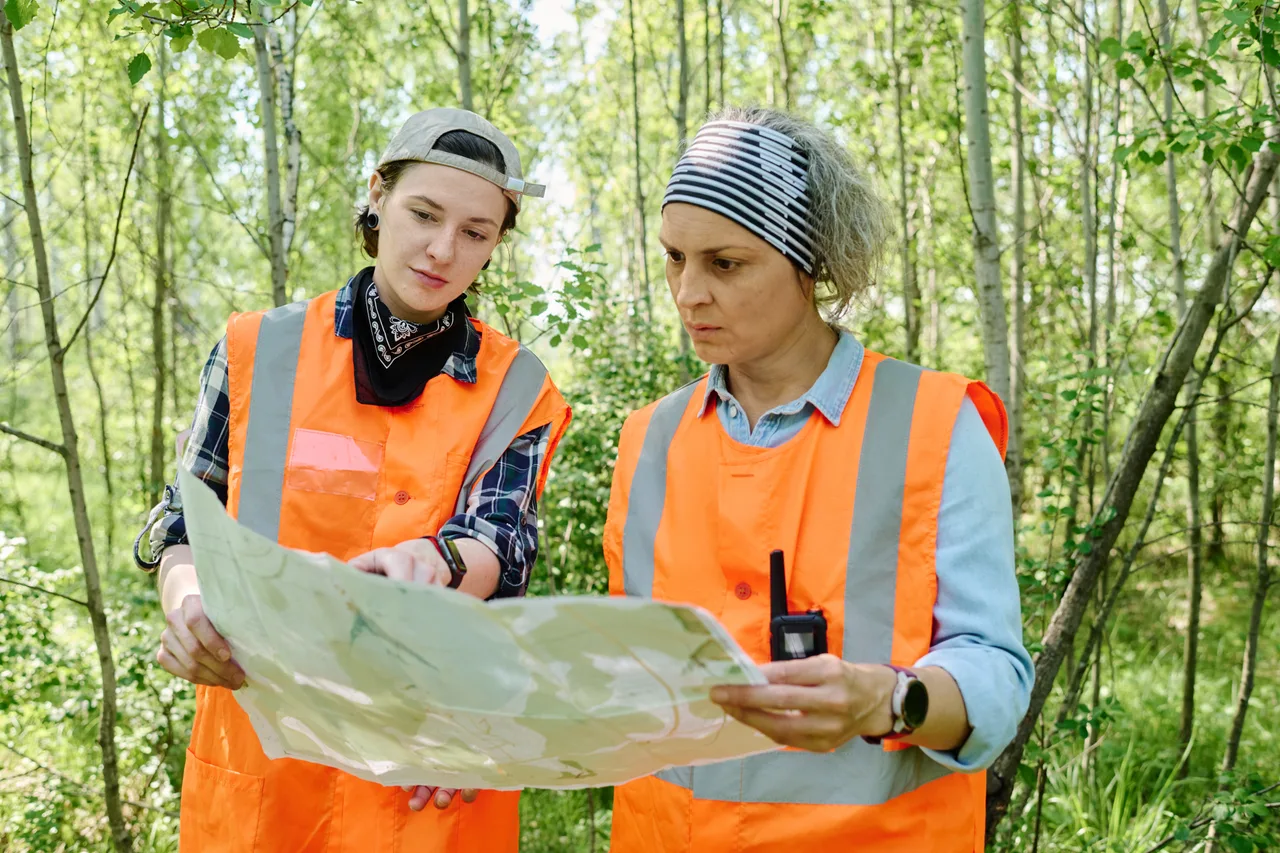 Two field surveyors in safety vests reviewing a map in a forest