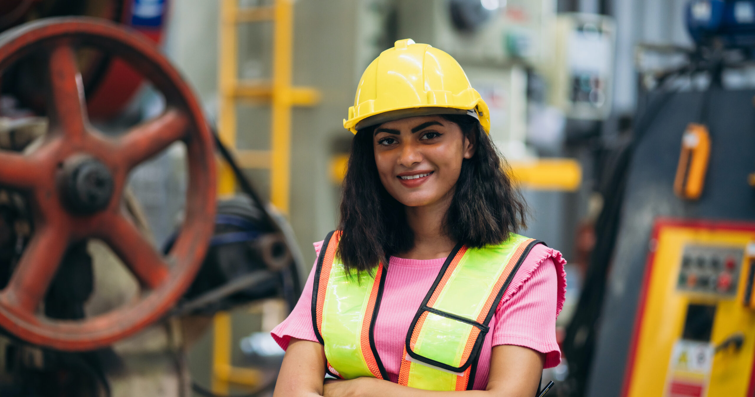 An engineer in a yellow hard hat and safety vest standing with folded arms beside plant equipment