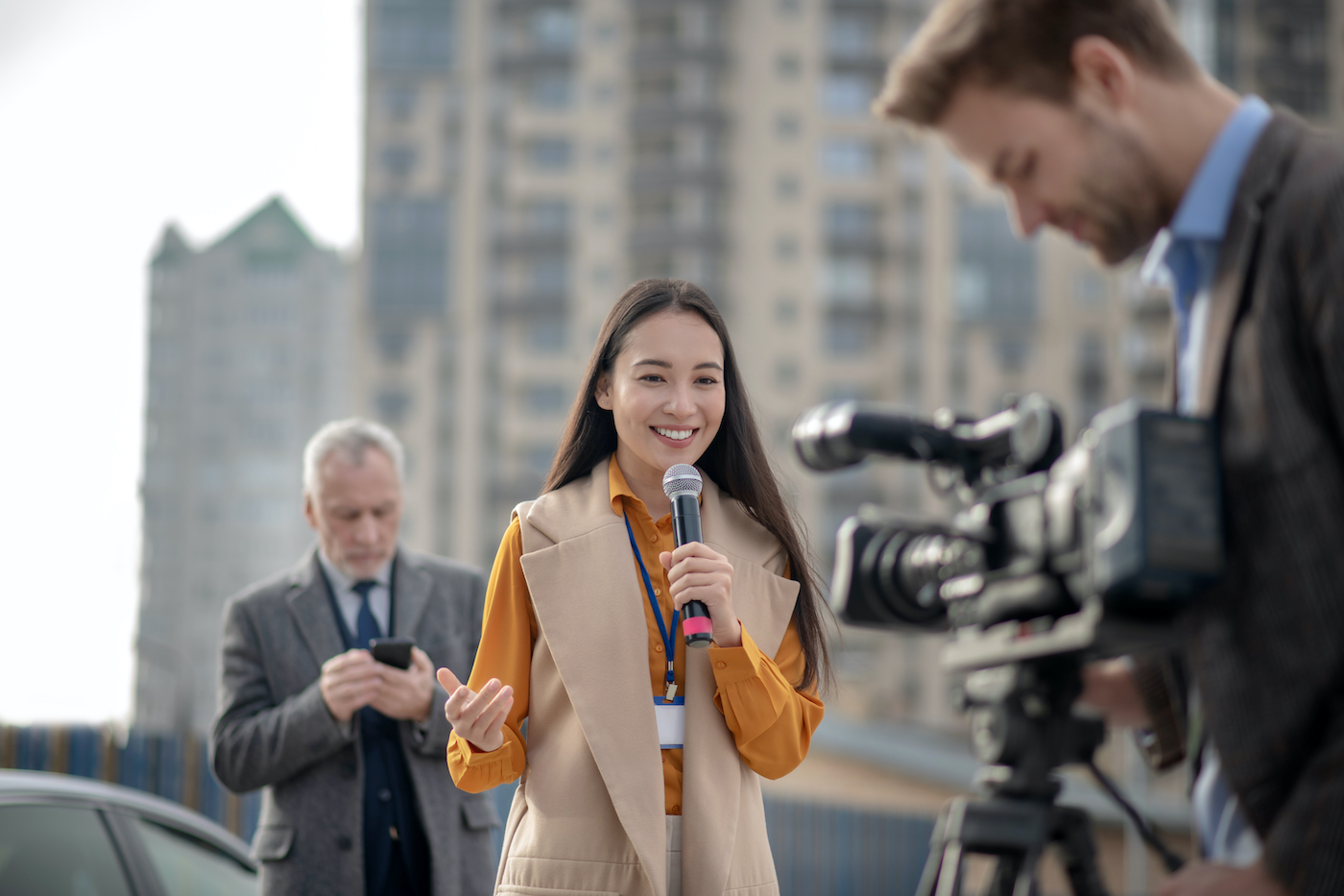 A reporter with a microphone speaking to a camera operator in the street while a man checks his phone behind her