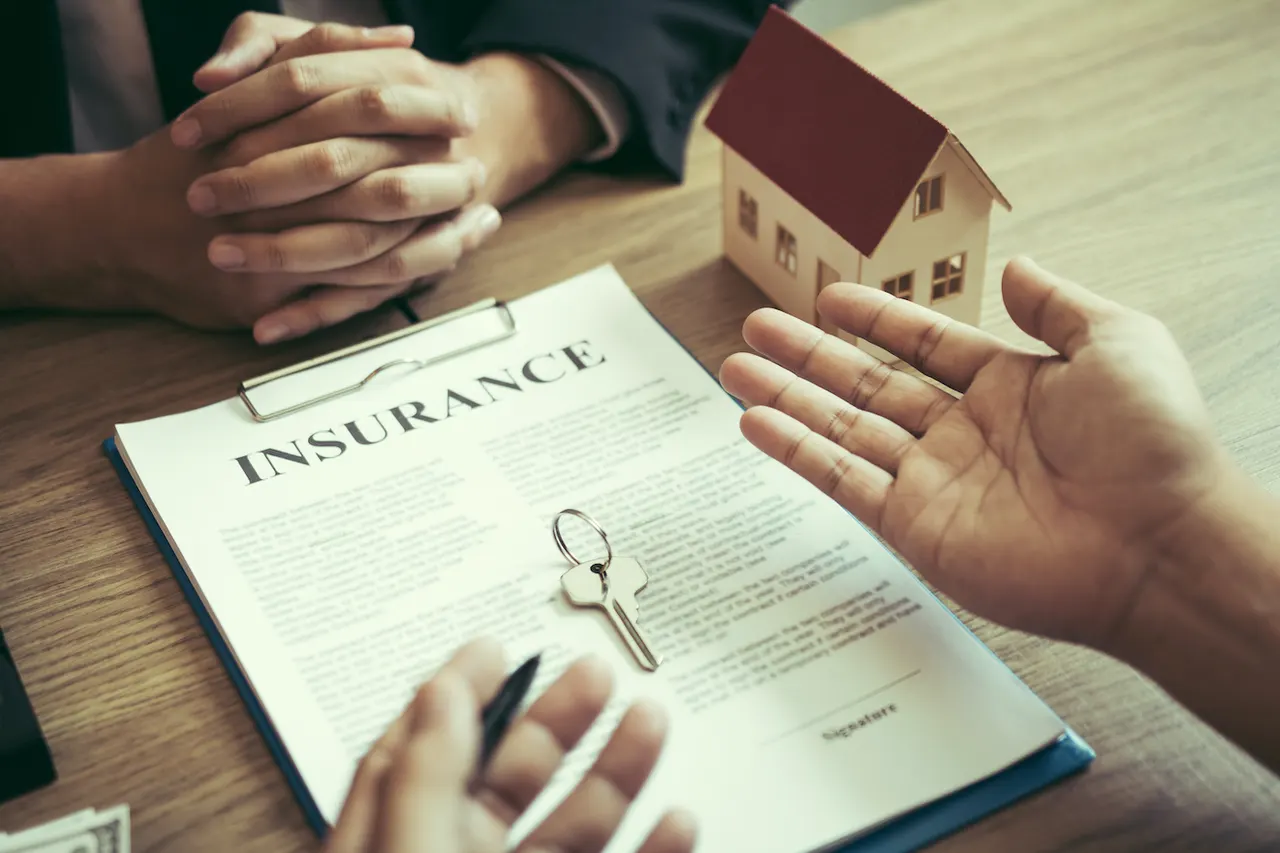 Two people across a desk with a printed insurance document, a model house and a set of keys