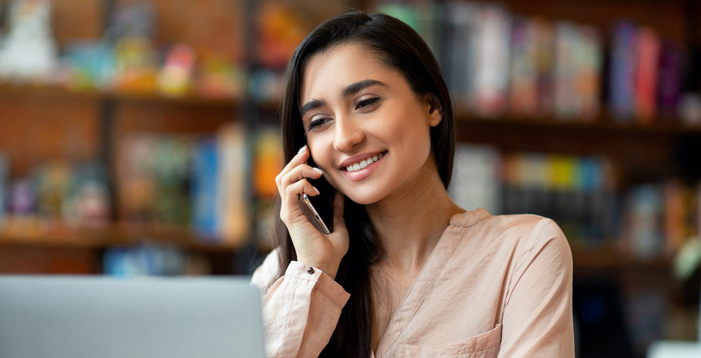 A woman smiling on a phone call at a laptop in front of bookshelves
