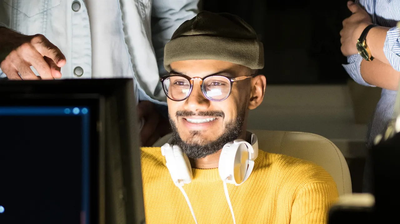 A smiling man in glasses with headphones round his neck working at a screen late at night