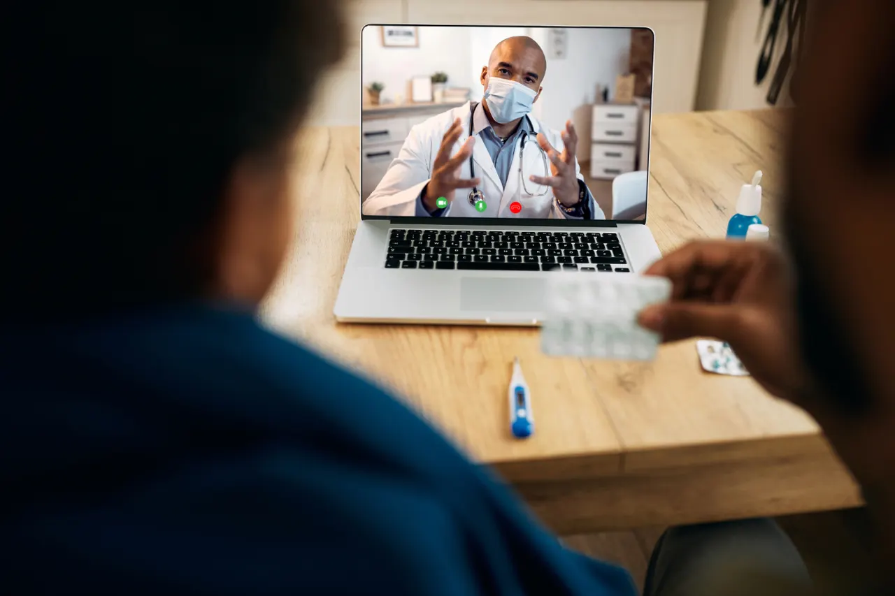 A person on a video call with a masked doctor shown on a laptop screen, holding a blister pack of tablets