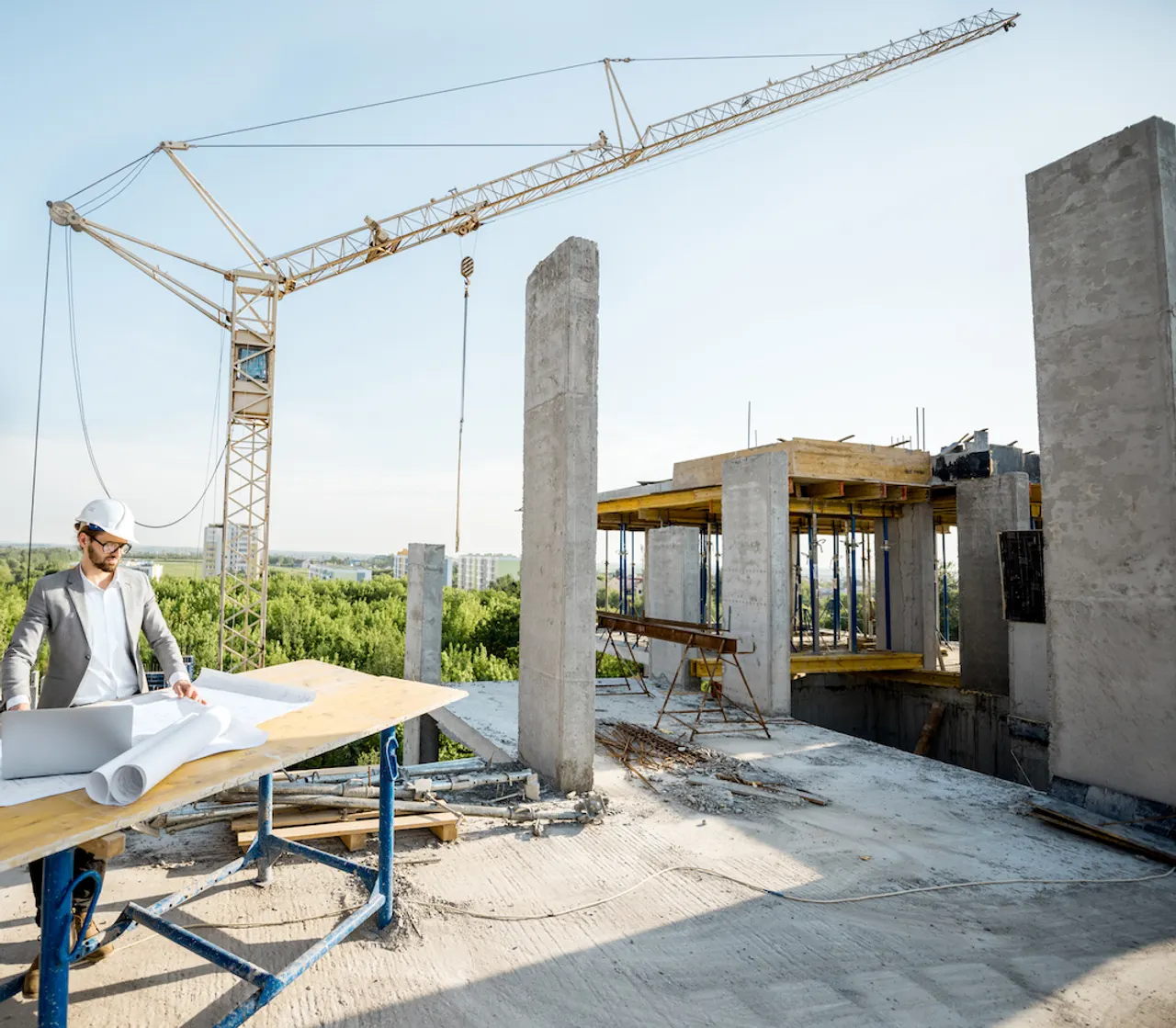 A site engineer in a hard hat reviewing plans and a laptop on a table at a concrete-frame building site, a tower crane overhead