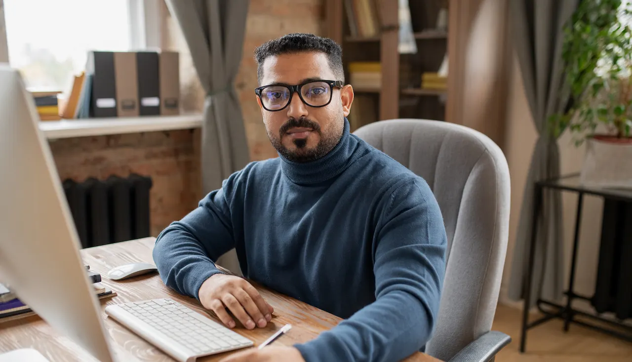 A man in glasses and a dark jumper seated at a desk with a desktop computer and keyboard