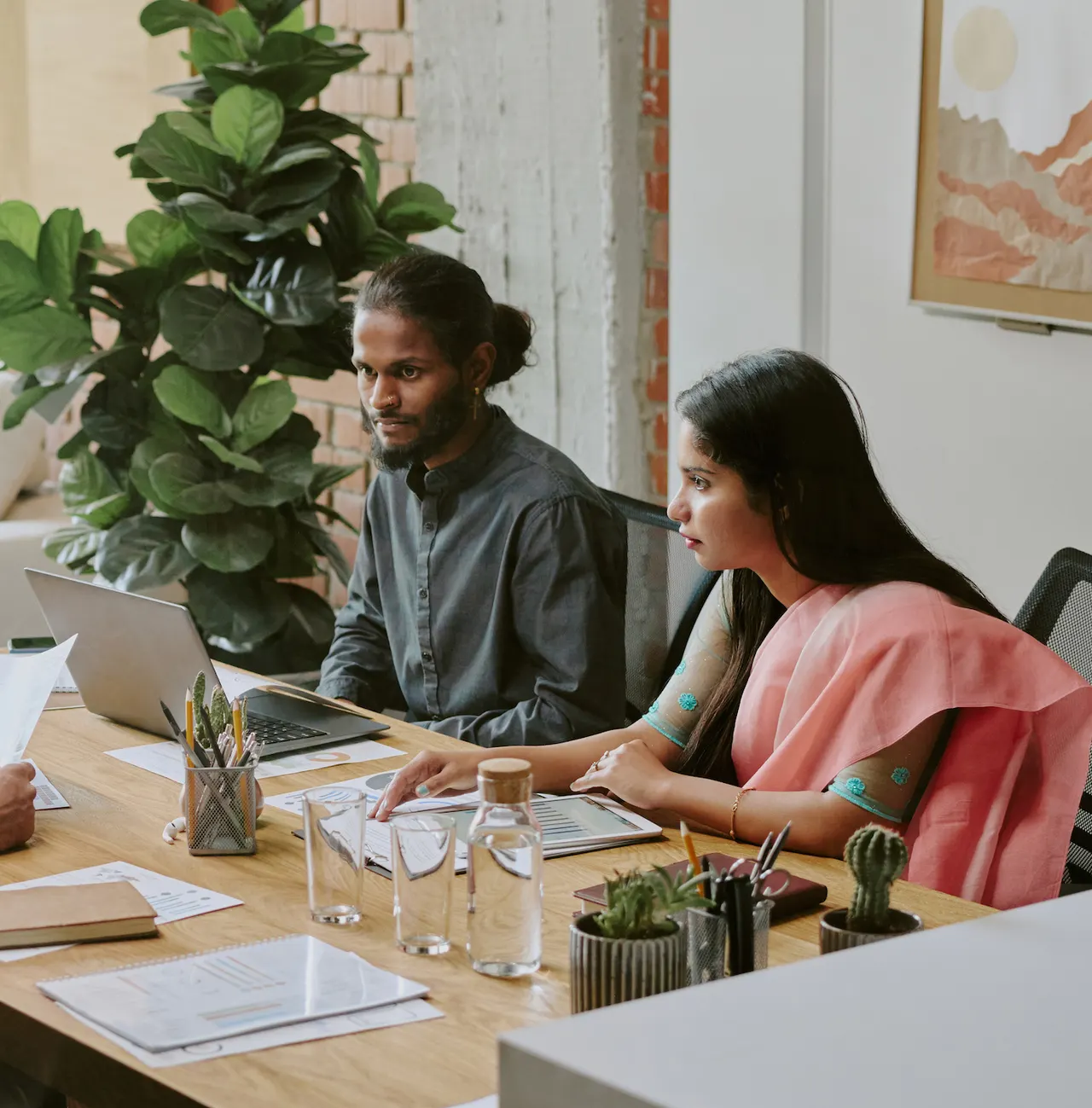 Two colleagues at a meeting table with a laptop and papers, plants behind them