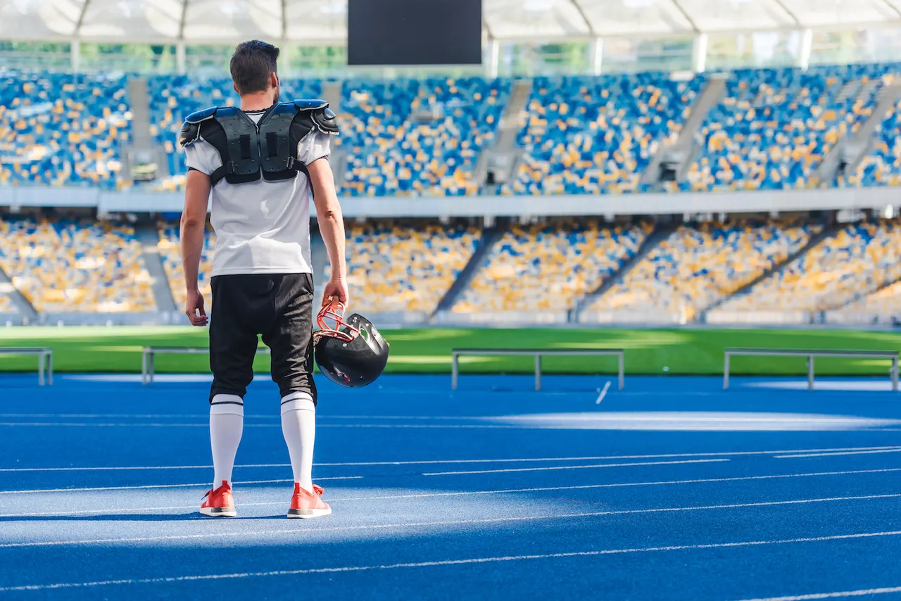 A player in shoulder pads holding his helmet, seen from behind on the running track of an empty stadium