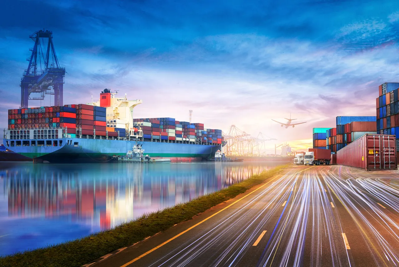 A loaded container ship beside a quay crane, with container stacks and trucks on a road along the waterfront