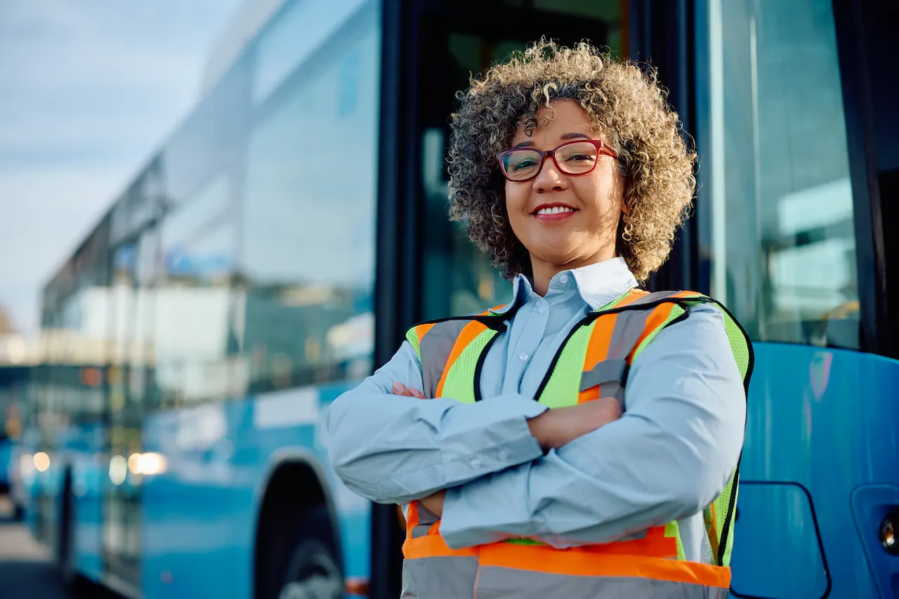 A transit worker in a high-visibility vest with arms crossed, standing at the door of a bus