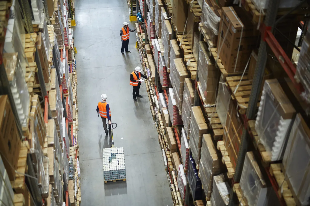 Warehouse staff in hard hats and high-visibility vests in a tall racked aisle, seen from above, one pulling a pallet truck