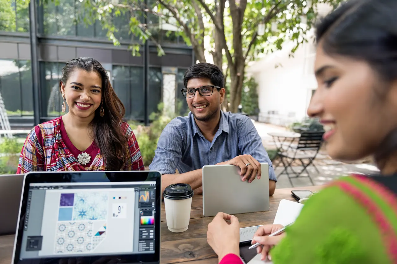 Three colleagues at an outdoor table, a laptop in front of them showing a pattern in a design application
