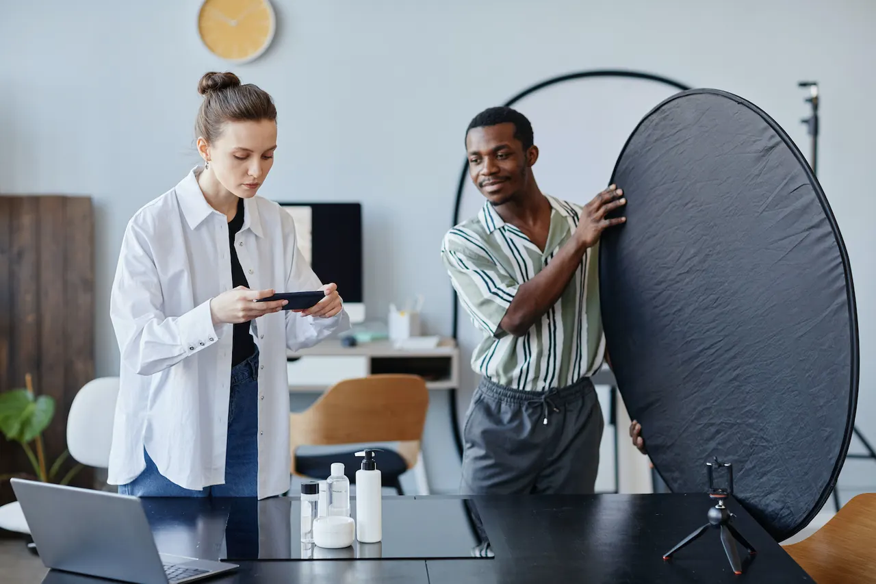 A photographer framing plain product bottles with a phone while a colleague holds up a large reflector