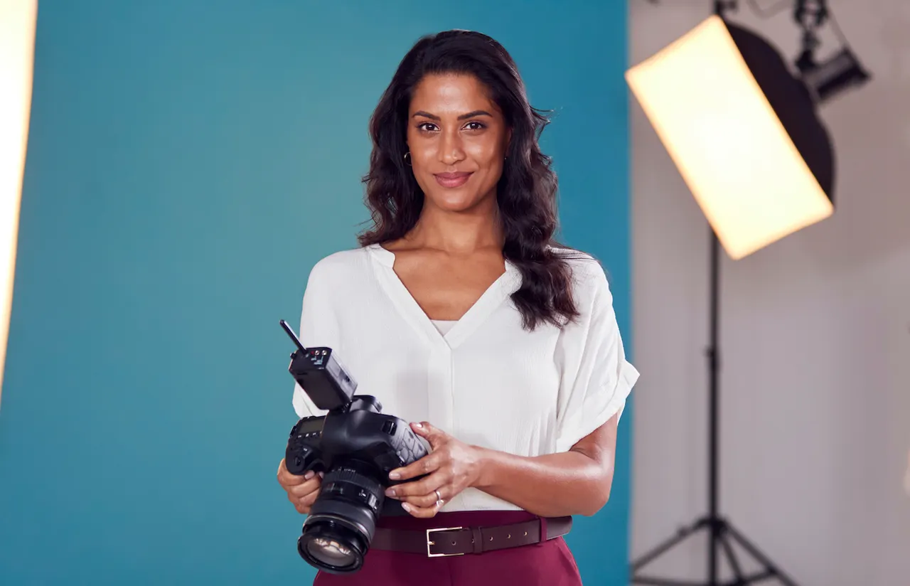 A photographer holding a camera fitted with a wireless trigger in front of a studio backdrop and softbox