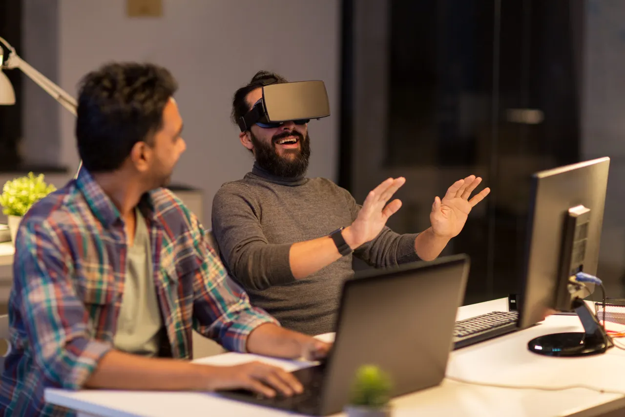 A developer in a virtual reality headset reaching out with both hands while a colleague watches from a laptop