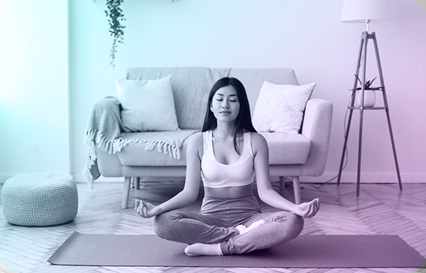A woman sitting cross-legged on an exercise mat in a living room, eyes closed in meditation
