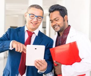 Young Indian doctor with businessman talking in hospital hallway - Salesman holding pad showing health care equipment at asian medical director - Concept of sales business deals with multiracial men